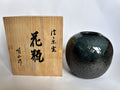 A dark glazed ceramic vase with gold floral details stands next to its light wood presentation box with black calligraphy, photographed against a plain white background.