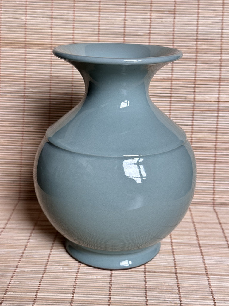 A glossy ceramic vase with a rounded body and flared rim, finished in a pale blue-green glaze, set against a woven straw mat background.