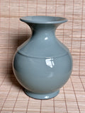 A glossy ceramic vase with a rounded body and flared rim, finished in a pale blue-green glaze, set against a woven straw mat background.