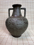 A dark brown ceramic vase with a bulbous body, two side handles, and geometric relief patterns, set against a woven straw mat background.
