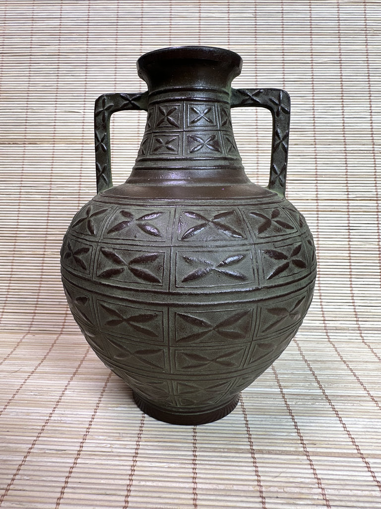 A dark brown ceramic vase with a bulbous body, two side handles, and geometric relief patterns, set against a woven straw mat background.