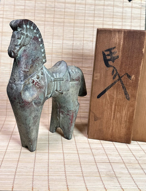 An ancient-style bronze horse figurine with a weathered greenish patina, displayed on a bamboo mat next to a wooden block with a black painted symbol.