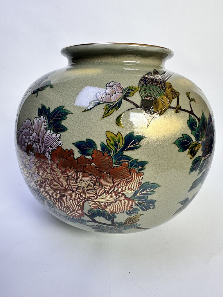 A round ceramic vase with a glossy, pale gold-beige glaze decorated with hand-painted floral and bird motifs, including a large pinkish-brown flower and a green bird, set against a plain white background.