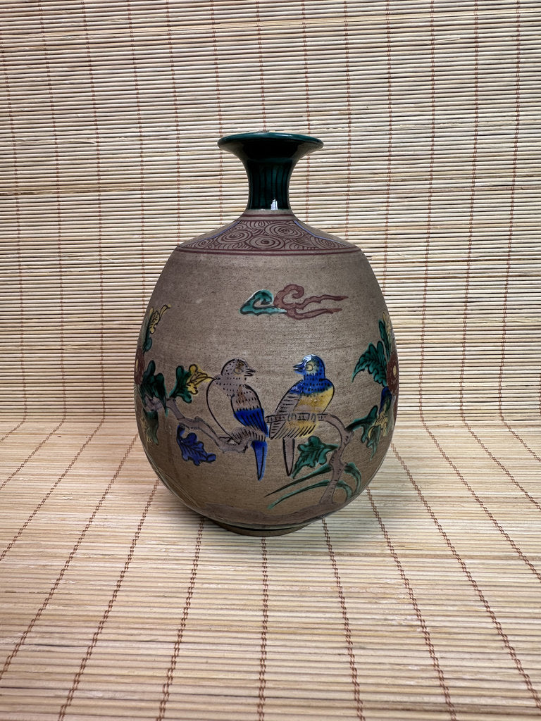 A vintage ceramic vase with a bulbous body, a narrow dark green neck, and hand-painted bird and cloud motifs in blue, green, and yellow on a beige background, set against a bamboo mat backdrop.