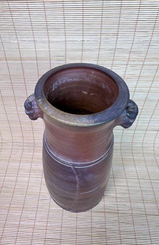 A rustic brown ceramic vase with a wide, flared rim and two small side handles, photographed from above on a woven straw mat.