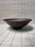 A dark brown ceramic bowl with a matte, textured glaze, placed on a light beige bamboo mat against a matching bamboo background.