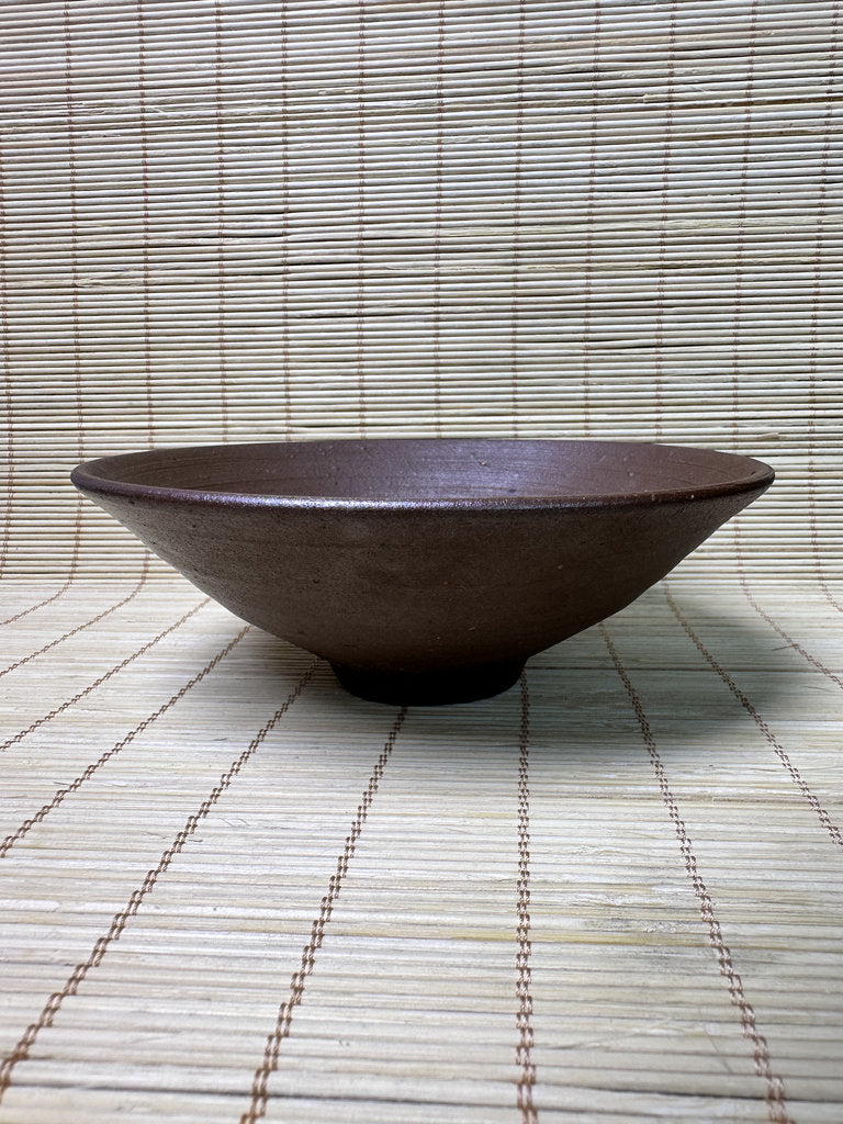 A dark brown ceramic bowl with a matte, textured glaze, placed on a light beige bamboo mat against a matching bamboo background.