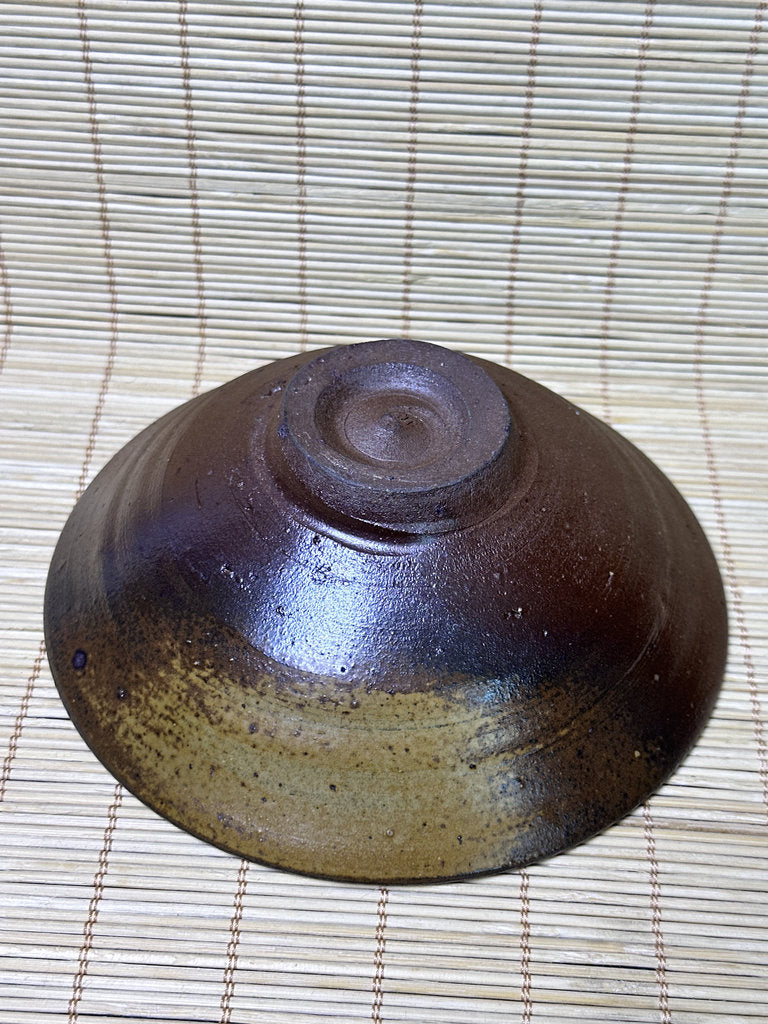 A handmade ceramic bowl with a dark brown and black glaze, shown upside down on a light-colored bamboo mat.