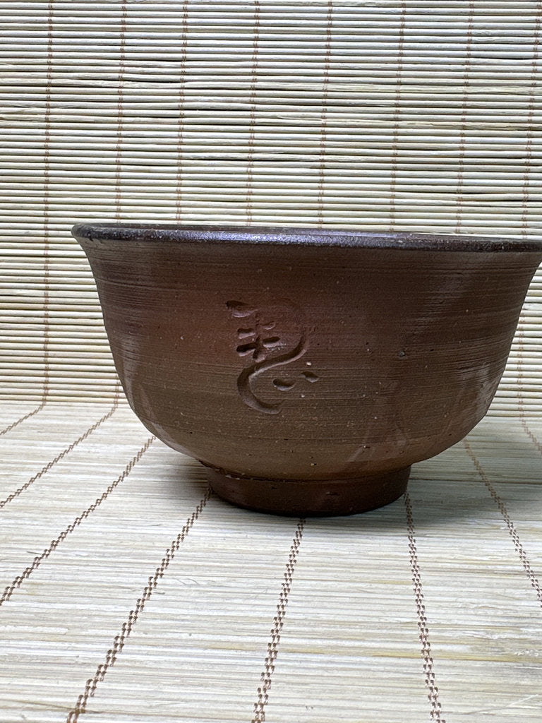 A dark brown handmade ceramic bowl with a textured glaze and an embossed Japanese character, set against a bamboo mat background.