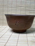 A dark brown handmade ceramic bowl with a textured glaze and an embossed Japanese character, set against a bamboo mat background.