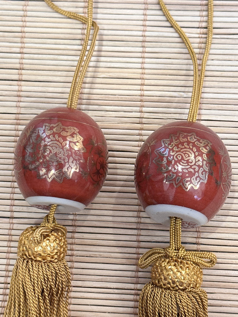 Two red glazed ceramic pendant ornaments with gold floral patterns and gold tassels, displayed on a light bamboo mat.