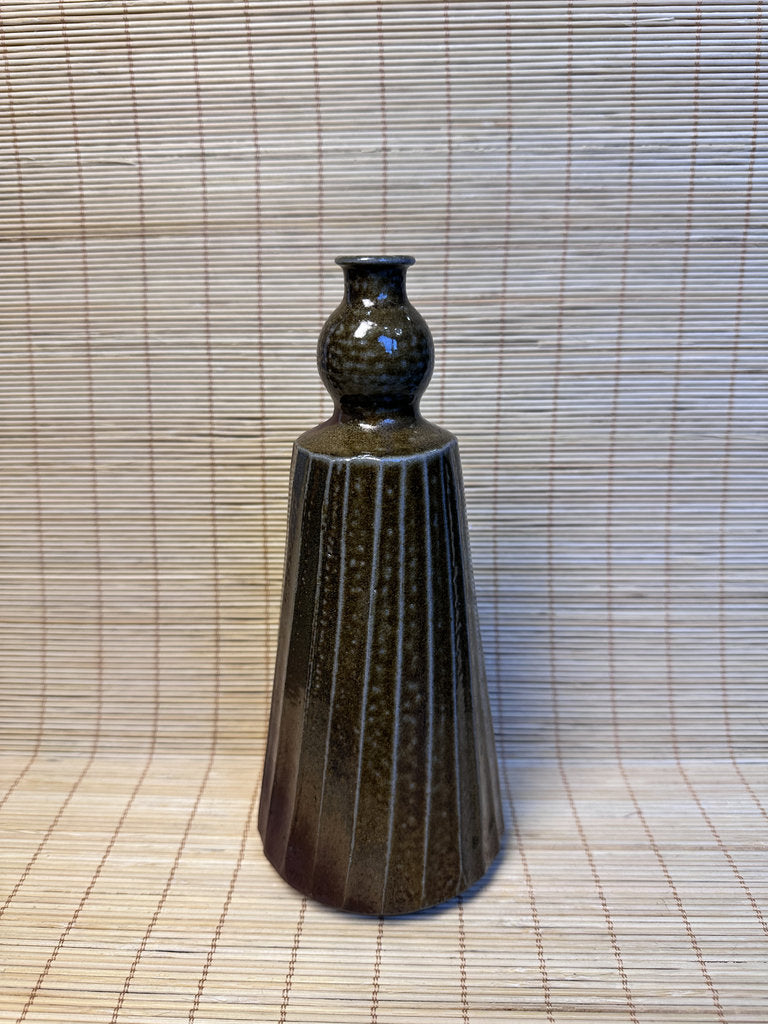 A dark brown ceramic vase with a textured, ribbed body and a bulbous neck, standing on a woven straw mat against a matching woven backdrop.