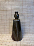 A dark brown ceramic vase with a textured, ribbed body and a bulbous neck, standing on a woven straw mat against a matching woven backdrop.