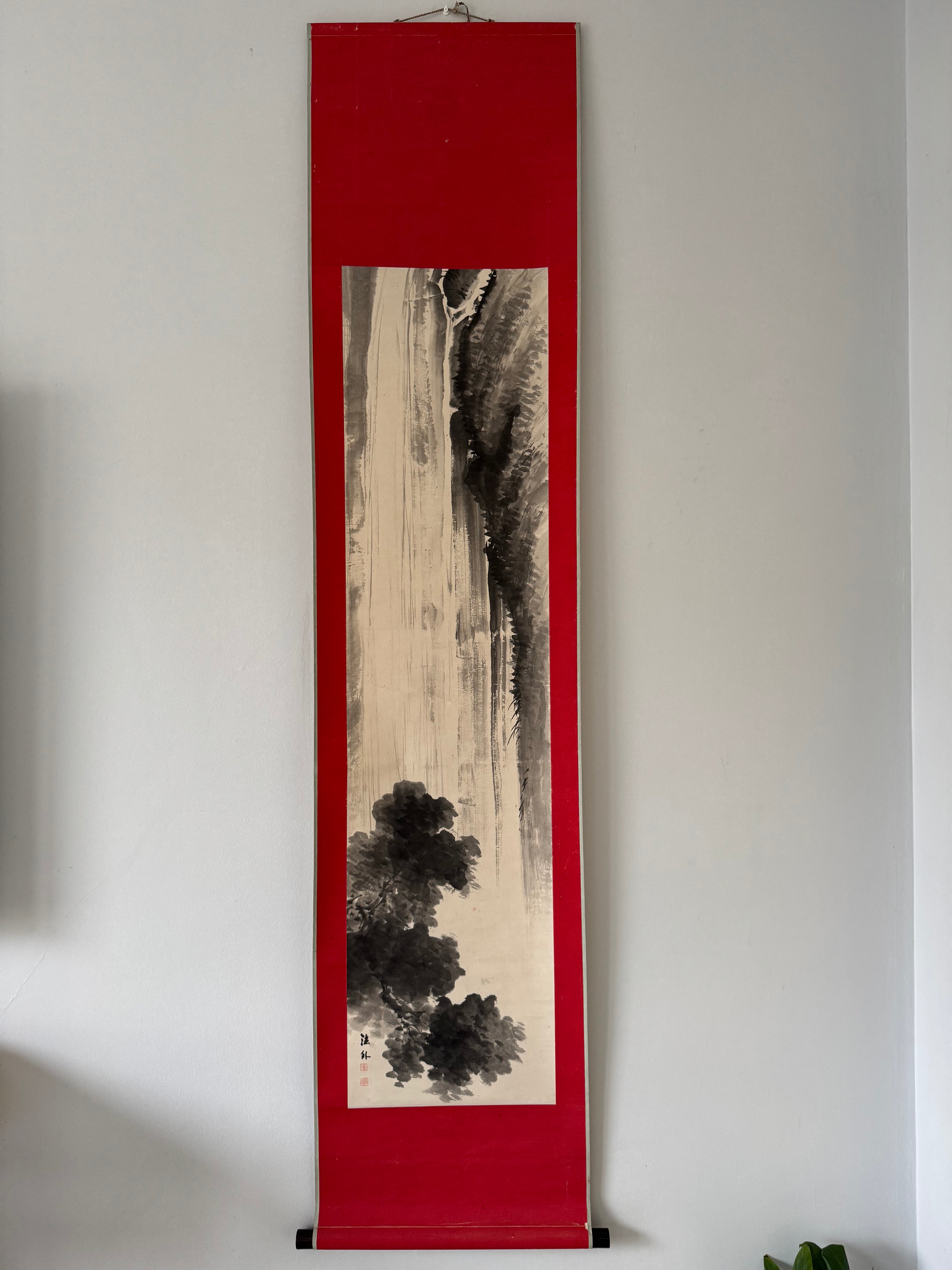 Vertical artwork with black ink design on beige paper, framed in red, against a white wall.