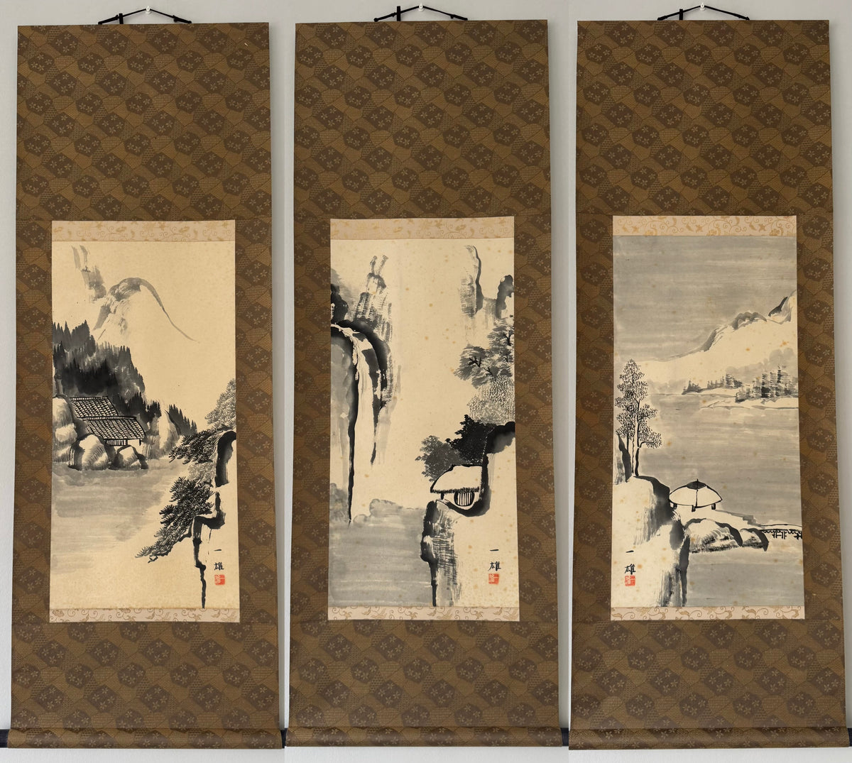 A three-panel traditional Chinese landscape wall scroll set, featuring ink wash paintings of a bird, a mountain scene, and a lakeside pavilion on beige backgrounds with brown patterned borders.