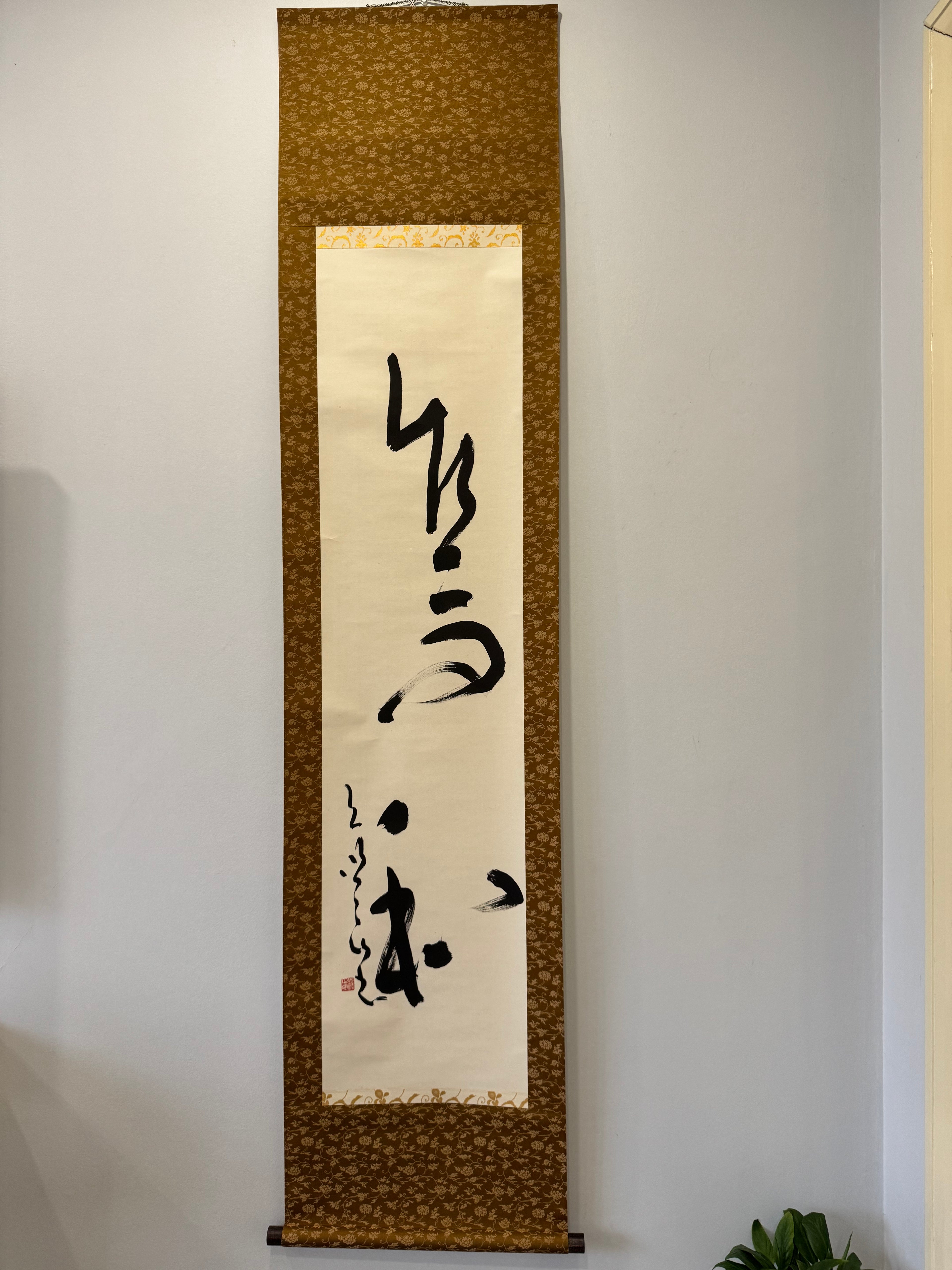 Vertical scroll with black calligraphy on beige paper against a white wall.