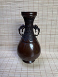 A dark brown glazed ceramic vase with a wide bulbous body, a flared neck, and two sculpted elephant ears on the sides, set against a woven straw mat background.