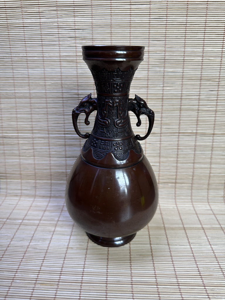 A dark brown glazed ceramic vase with a wide bulbous body, a flared neck, and two sculpted elephant ears on the sides, set against a woven straw mat background.