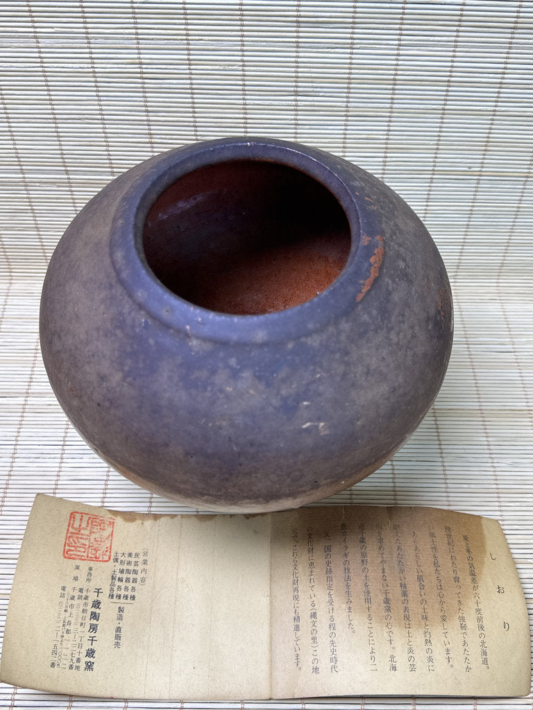 A round, open ceramic jar with a dark purple-brown glazed exterior and a reddish-brown interior, displayed on a beige woven mat with an old Japanese paper document underneath.