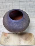 A round, open ceramic jar with a dark purple-brown glazed exterior and a reddish-brown interior, displayed on a beige woven mat with an old Japanese paper document underneath.