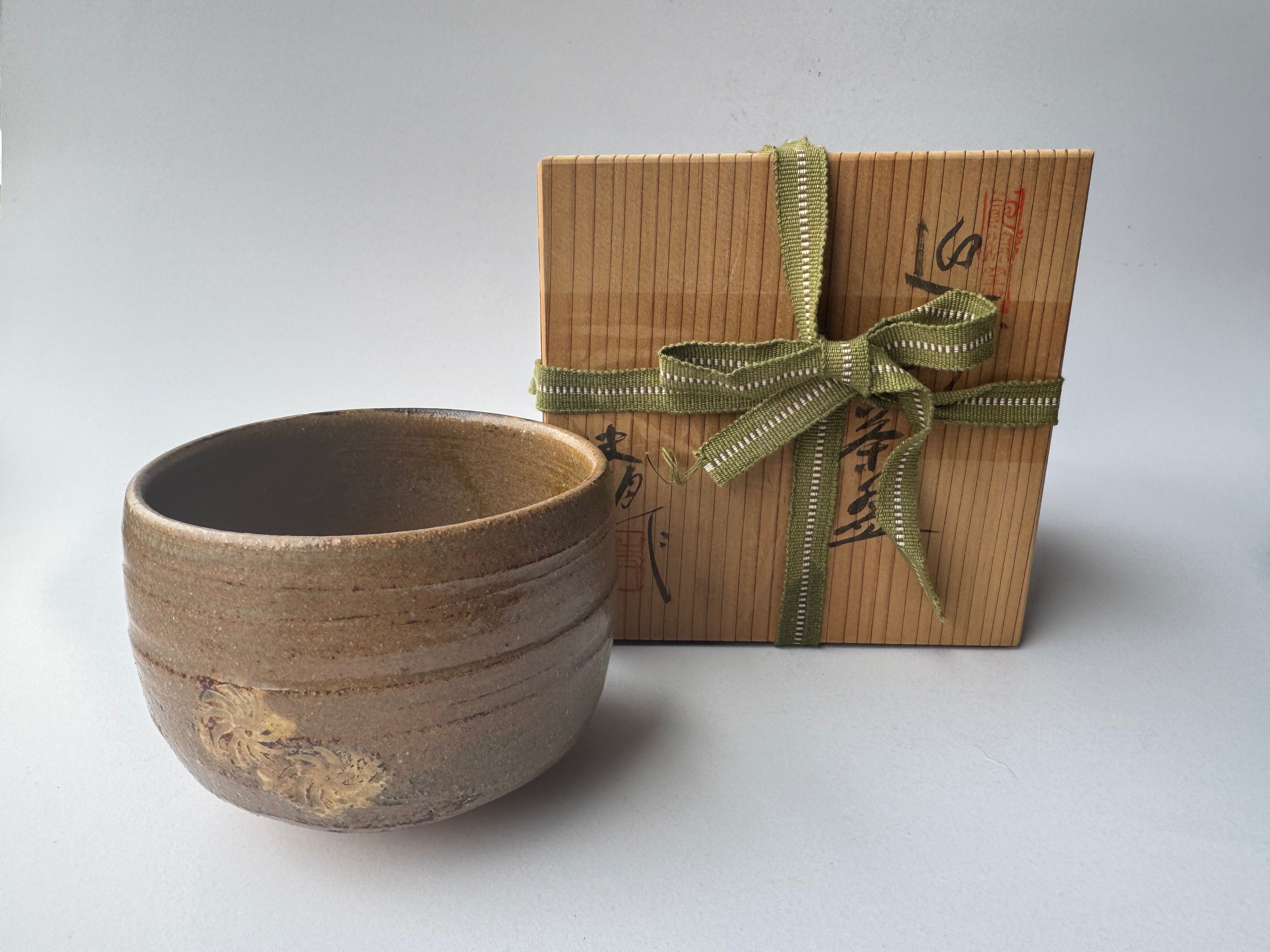 Ceramic bowl with gold patterns next to a gift box with a green ribbon on a white background