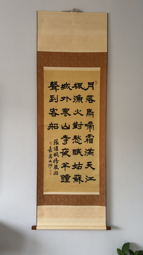 A traditional Jpanese calligraphy scroll hanging on a light gray wall. The scroll features black calligraphy characters on a beige paper background, bordered by a brown frame with beige hanging bands at the top and bottom.