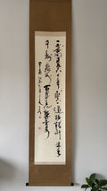 A traditional Japanese calligraphy scroll with black ink calligraphy on an off-white paper center, mounted on a brown wooden frame, hanging on a light gray wall.