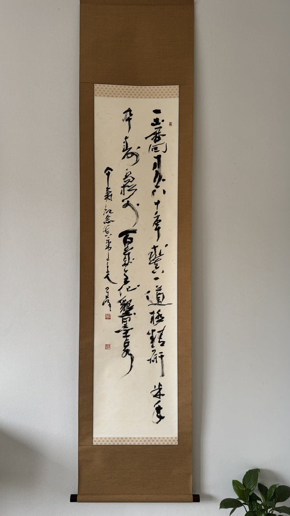 A traditional Japanese calligraphy scroll with black ink calligraphy on an off-white paper center, mounted on a brown wooden frame, hanging on a light gray wall.