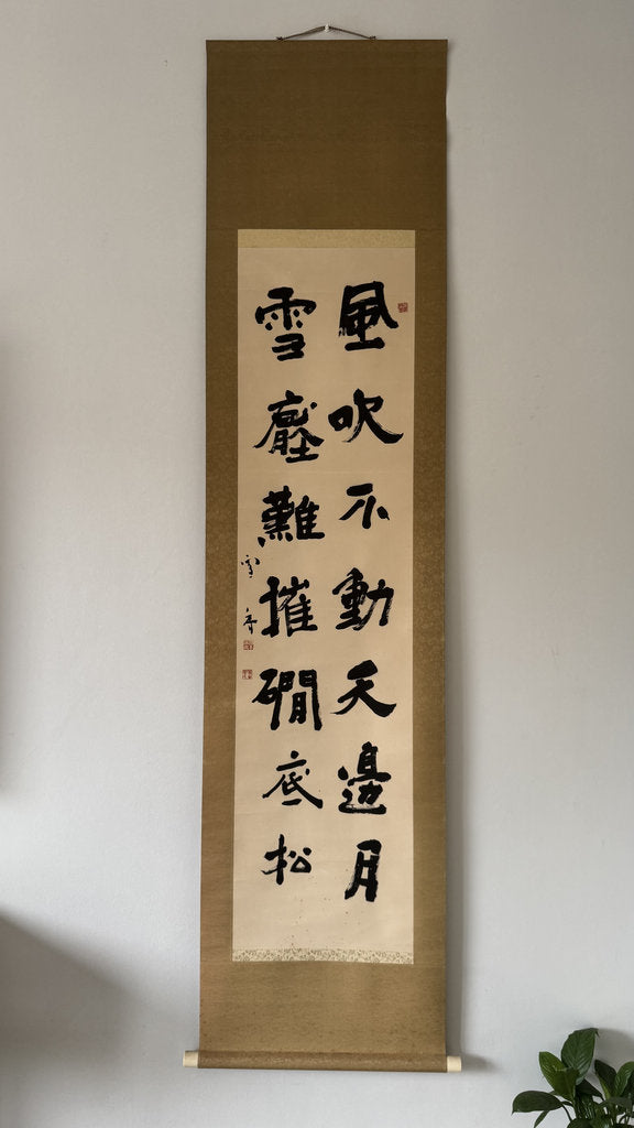 A traditional Japanese calligraphy scroll hanging on a light gray wall, featuring black ink characters on beige paper with a brown border, with a small green plant visible in the bottom right corner.