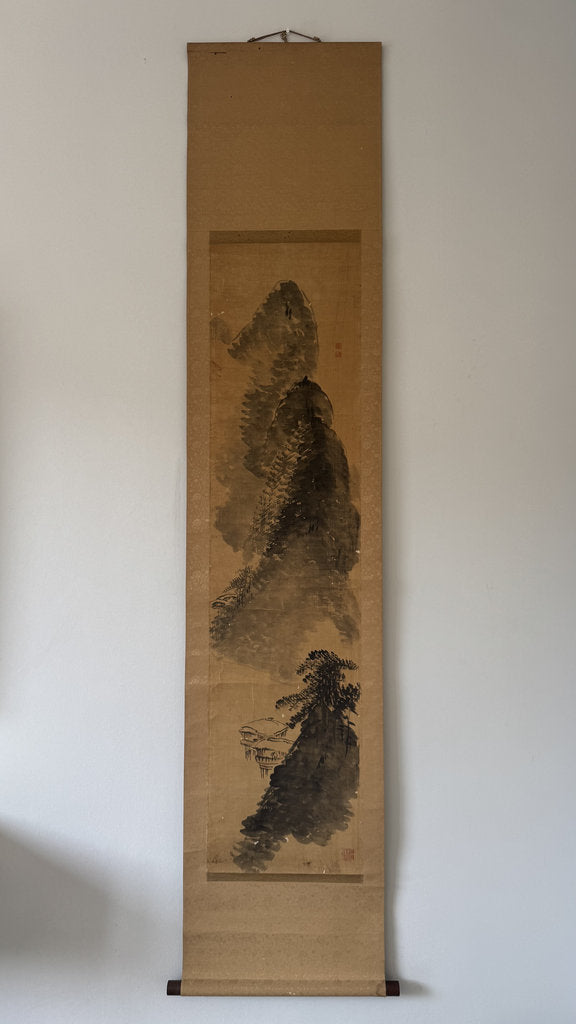 A traditional Japanese ink scroll painting featuring a mountain and tree scene, mounted on a light beige wall.