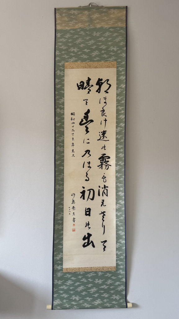 A traditional Japanese hanging calligraphy scroll with black brushstrokes on a beige paper center panel, bordered by a green patterned fabric and a light wood hanging rod against a plain wall.