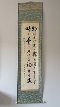 A traditional Japanese hanging calligraphy scroll with black brushstrokes on a beige paper center panel, bordered by a green patterned fabric and a light wood hanging rod against a plain wall.