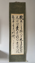 A traditional Japanese hanging scroll with black calligraphy on a beige paper center panel, mounted on a dark textured green fabric border, hanging against a plain light gray wall.