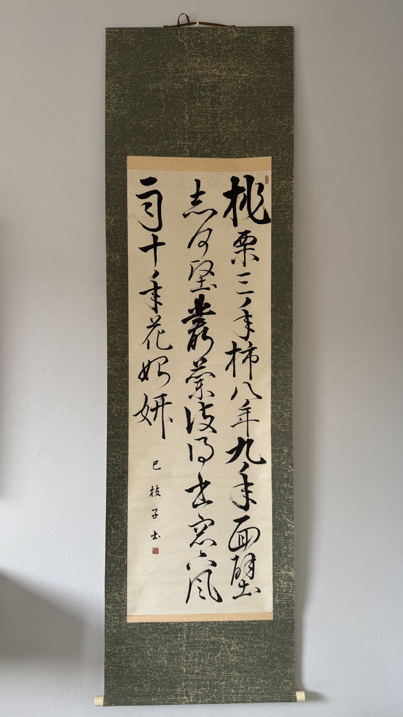 A traditional Japanese hanging scroll with black calligraphy on a beige paper center panel, mounted on a dark textured green fabric border, hanging against a plain light gray wall.