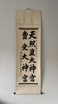 A traditional Japanese hanging calligraphy scroll with black brush-stroke characters on aged beige paper, mounted against a plain light gray wall.