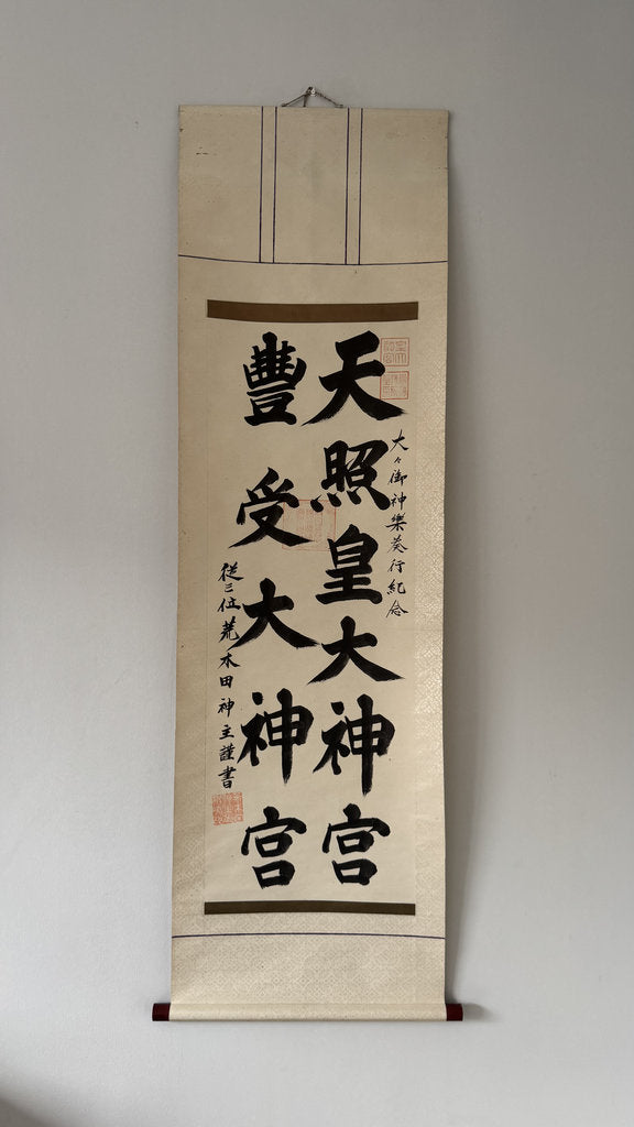 A traditional Japanese hanging calligraphy scroll with black brush-stroke characters on aged beige paper, mounted against a plain light gray wall.
