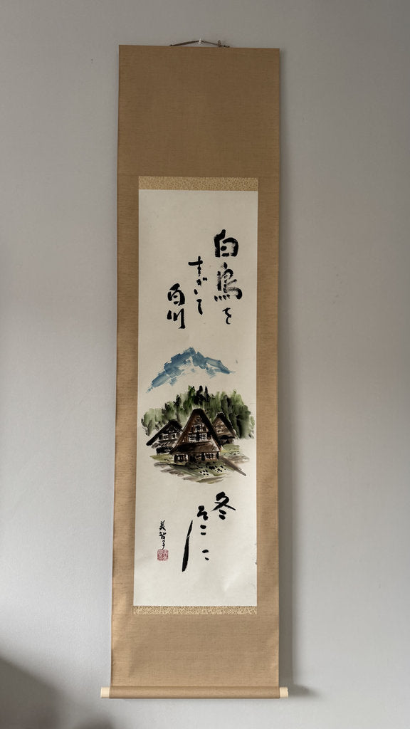 A traditional Japanese hanging scroll (kakemono) featuring a hand-painted winter landscape with mountains, houses, and black calligraphy on a white paper center panel, mounted on a beige paper border.