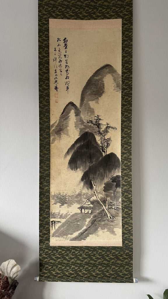 A traditional Japanes ink wash scroll painting hanging on a light gray wall. The scroll features a black ink landscape with mountains, trees, and a path, bordered by a dark patterned green fabric.