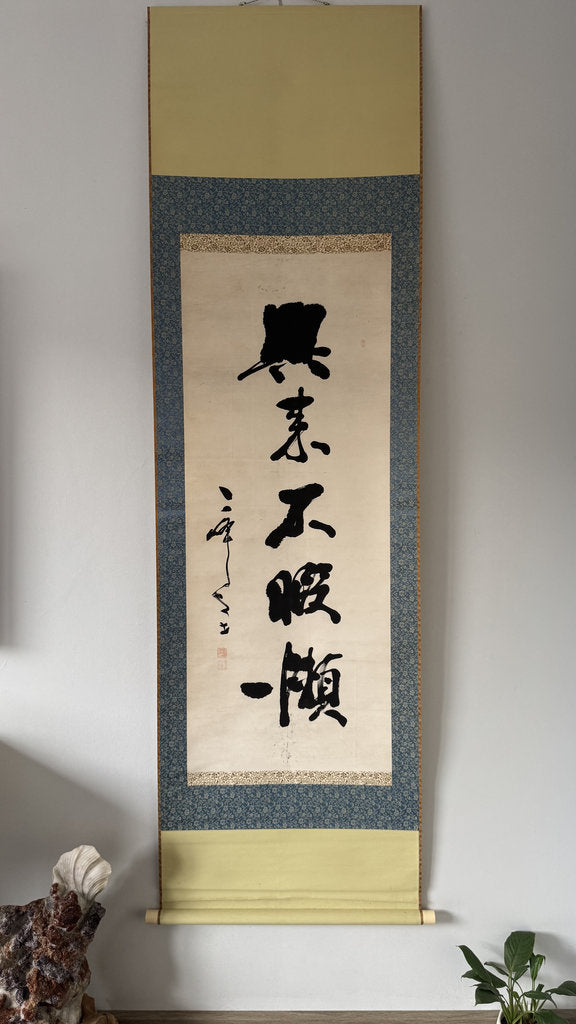 A traditional Japanese hanging scroll with black calligraphy on an off-white center panel, bordered by a blue patterned band and beige fabric ends, displayed against a light gray wall.