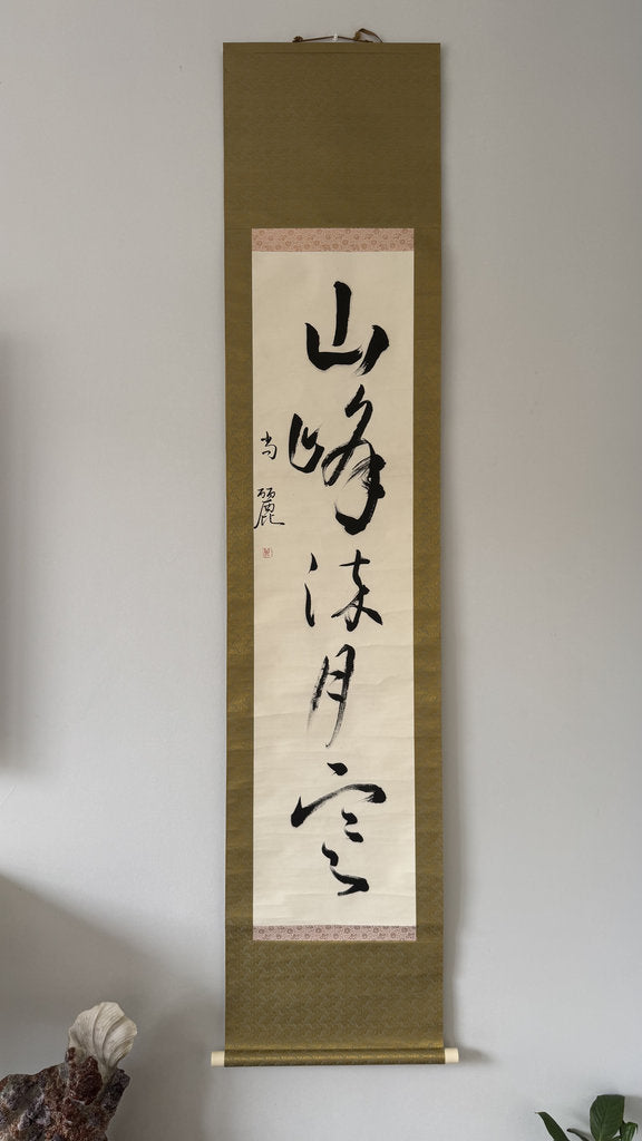 A traditional Japanese hanging scroll with black calligraphy on an off-white paper center, mounted on a textured olive-green fabric background, hanging on a light gray wall.