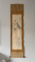 A traditional Japanese hanging scroll (kakemono) featuring black ink calligraphy and a landscape painting on aged beige paper, mounted on a brown wood frame and displayed against a neutral light gray wall.