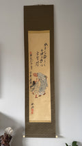A traditional Japanese hanging scroll (kakejiku) featuring ink calligraphy and a hand-painted illustration of an elderly man and child, mounted on a light gray wall.