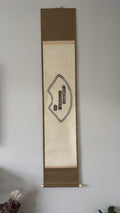 A traditional Japanese hanging scroll with a beige central panel, brown top and bottom borders, and black calligraphy, displayed on a light gray wall.