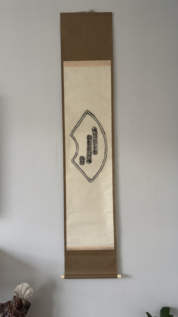 A traditional Japanese hanging scroll with a beige central panel, brown top and bottom borders, and black calligraphy, displayed on a light gray wall.