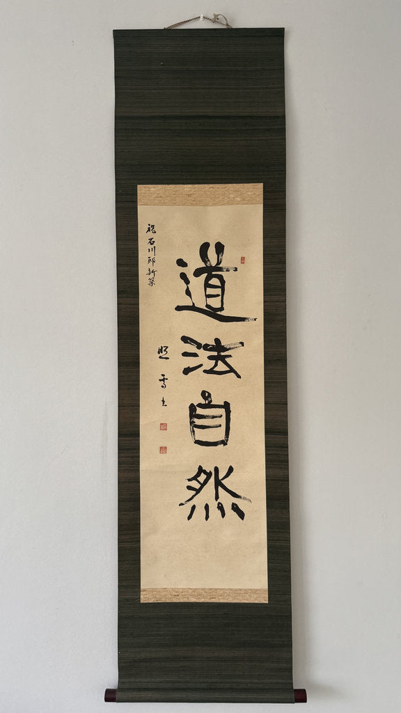 A vertical Japanese calligraphy scroll featuring bold black brushstrokes on aged beige paper, mounted against a dark wood background. The calligraphy displays the characters '武品小宝' with red seal stamps and calligraphic inscriptions on the left.