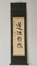 A vertical Japanese calligraphy scroll featuring bold black brushstrokes on aged beige paper, mounted against a dark wood background. The calligraphy displays the characters '武品小宝' with red seal stamps and calligraphic inscriptions on the left.