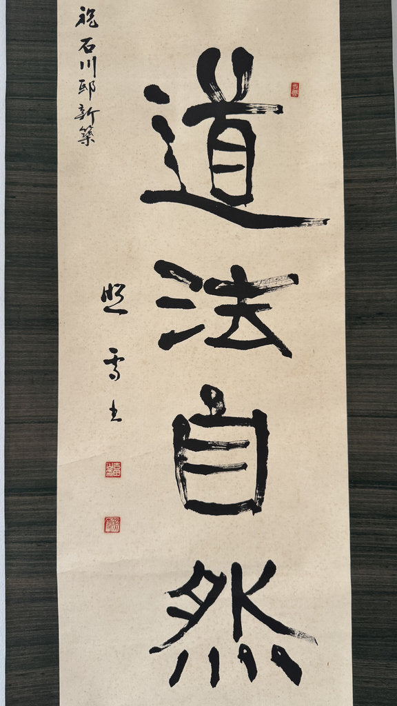 A vertical Japanese calligraphy scroll featuring bold black brushstrokes on aged beige paper, mounted against a dark wood background. The calligraphy displays the characters '武品小宝' with red seal stamps and calligraphic inscriptions on the left.