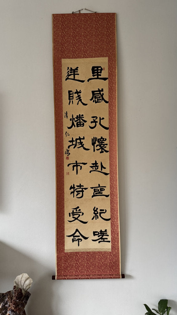 A vertical Japanese calligraphy scroll with black characters on a beige paper center, mounted on a textured reddish-brown fabric border, hanging on a plain white wall.