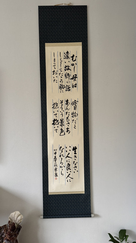 A vertical Japanese calligraphy scroll with black brushstrokes on a cream paper center panel, mounted on a dark textured navy blue hanging sleeve, shown hanging on a white wall.