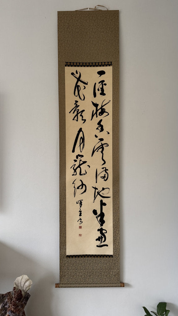 A vertical Japanese calligraphy scroll with black brushstrokes on aged beige paper, mounted on a textured brown and gray border, hanging on a plain light gray wall.